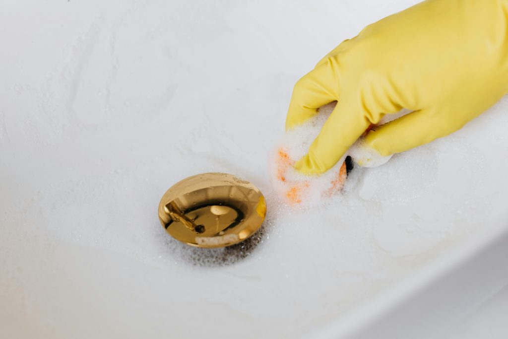 Close-up of a hand in a glove cleaning a foam-covered gold drain in a sink.